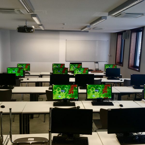 Foto: ein Seminarraum, der in Tischreihen aufgeteilt ist, auf jedem Tisch steht ein Computer. Sie sind alle frontal ausgerichtet, vorne im Raum ist ein Whiteboard und ein Beamer
